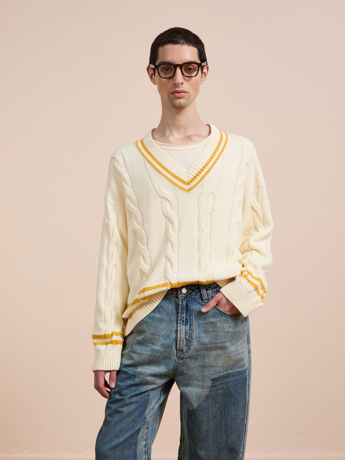 A person in FOUND's V-Neck Cricket Sweater—a cream unisex knit with yellow stripes—paired with blue jeans and glasses stands against a plain beige background.