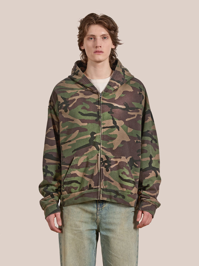 A person stands against a plain background wearing the Washed Camouflage Zip Hoodie by FOUND, paired with light blue jeans.