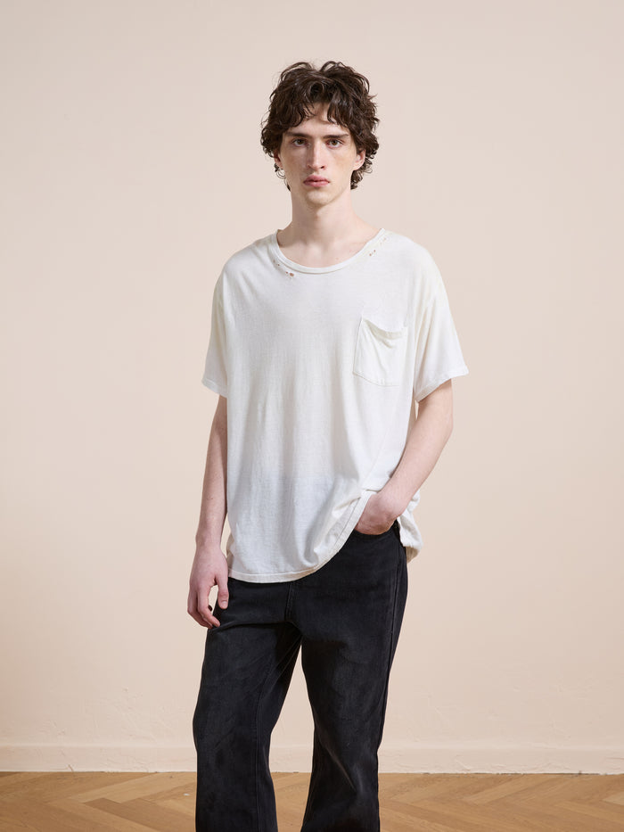 Indoors against a plain wall, a person wears FOUND’s Weathered Pocket Tee—crafted in soft cotton with vintage details and small holes—paired with dark pants and one hand casually slipped into a pocket.