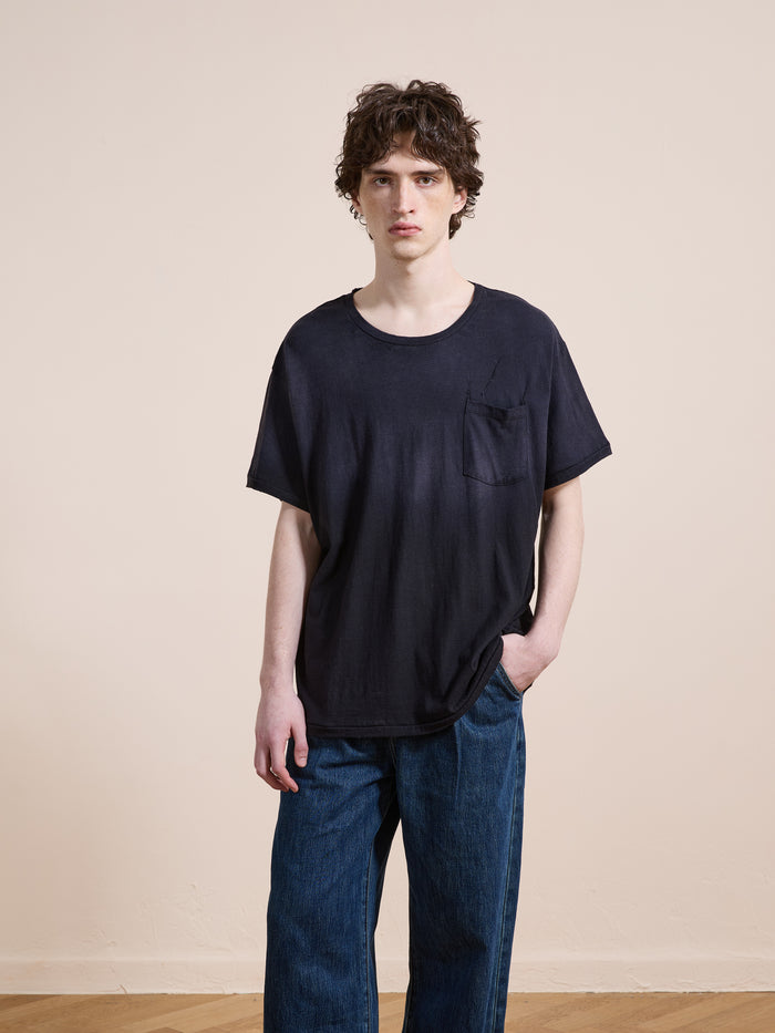 A person with wavy brown hair wears the FOUND Weathered Pocket Tee—a relaxed fit black t-shirt with a chest pocket—paired with blue jeans, standing indoors against a plain light-colored wall.