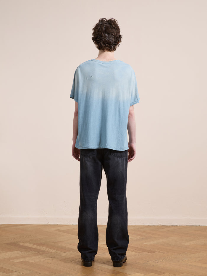 A person stands indoors facing away, wearing the FOUND Weathered Pocket Tee—a light blue unisex relaxed-fit shirt—paired with dark jeans on a wooden floor against a plain beige wall.