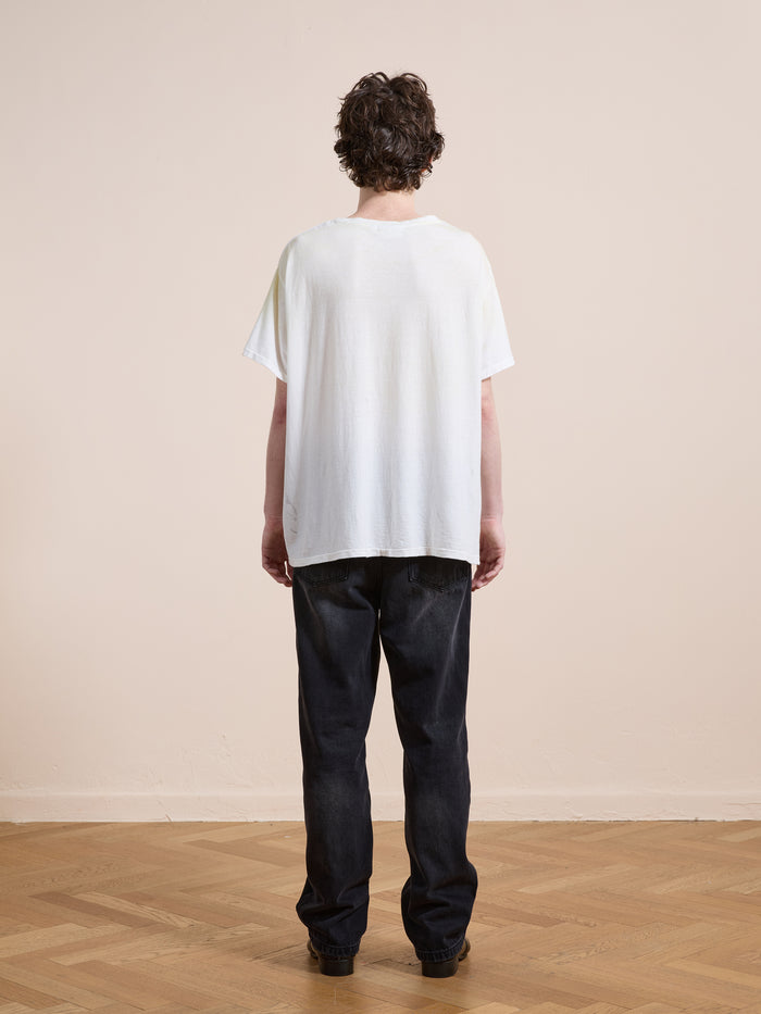 A person with short curly hair stands facing away, wearing the FOUND Weathered Pocket Tee in white with black jeans on a wooden floor against a plain beige wall.