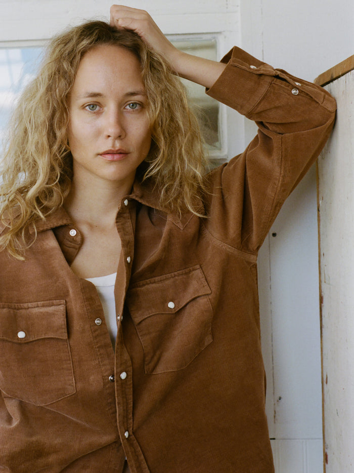 A person with long curly blonde hair wears the FOUND Western Corduroy Shirt in a unisex relaxed fit, standing indoors with one arm resting on a white wall and looking at the camera.