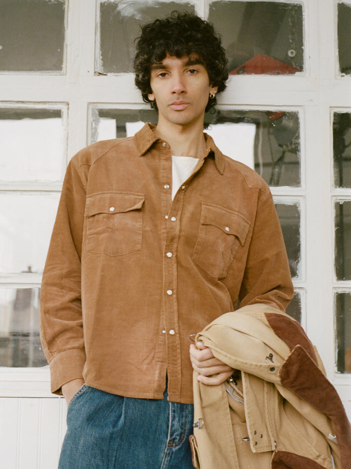 Wearing the FOUND Western Corduroy Shirt over a white top and blue jeans, a person with curly hair stands by a window holding a tan jacket.