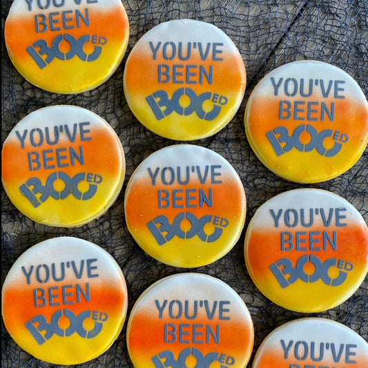 You've Been Booed Cookie Stencil shown on nine sugar cookies that have airbrushed backgrounds resembling candy corn with the stencil decorated in black royal icing and all laying on a black netted background
