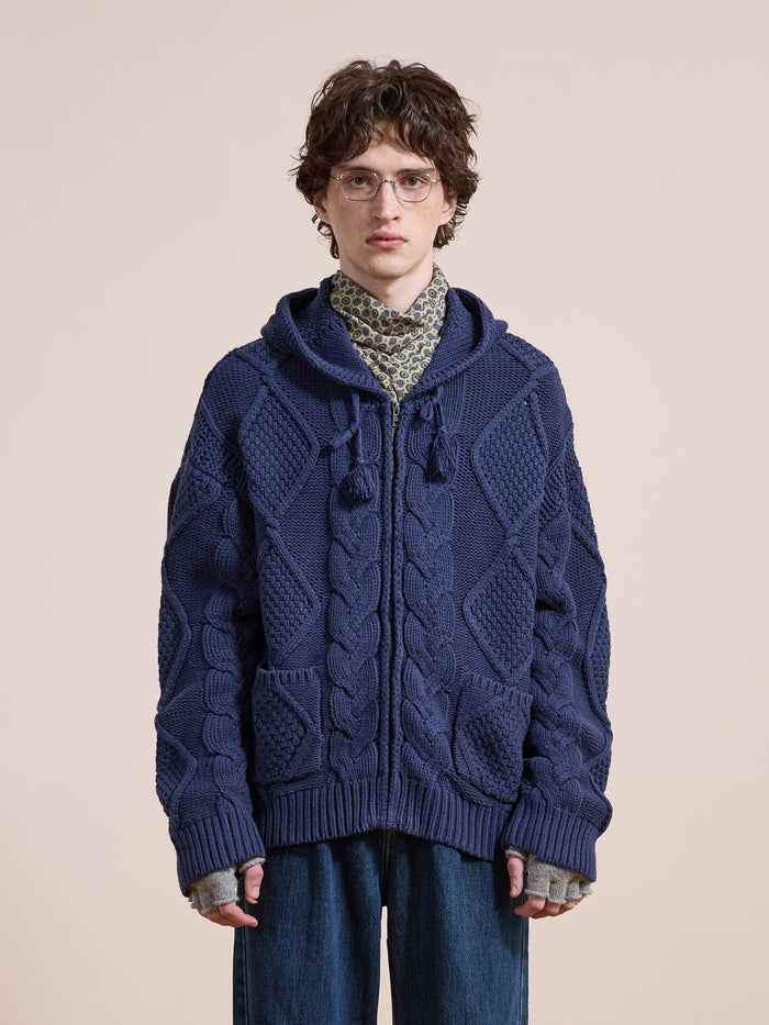 Wearing the Found Zip Up Cable Knit Hoodie in textured navy blue over a patterned shirt and loose denim jeans, a person with glasses stands against a plain beige background.