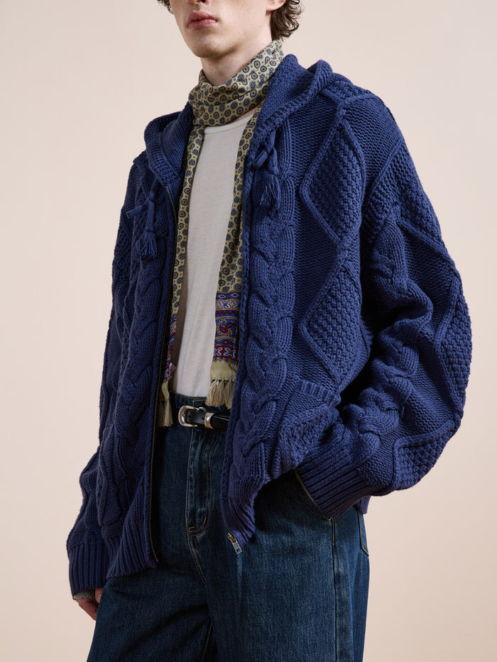 Someone wearing the Found Zip Up Cable Knit Hoodie in textured blue over a white shirt, with a patterned scarf and blue jeans, stands against a plain beige background.