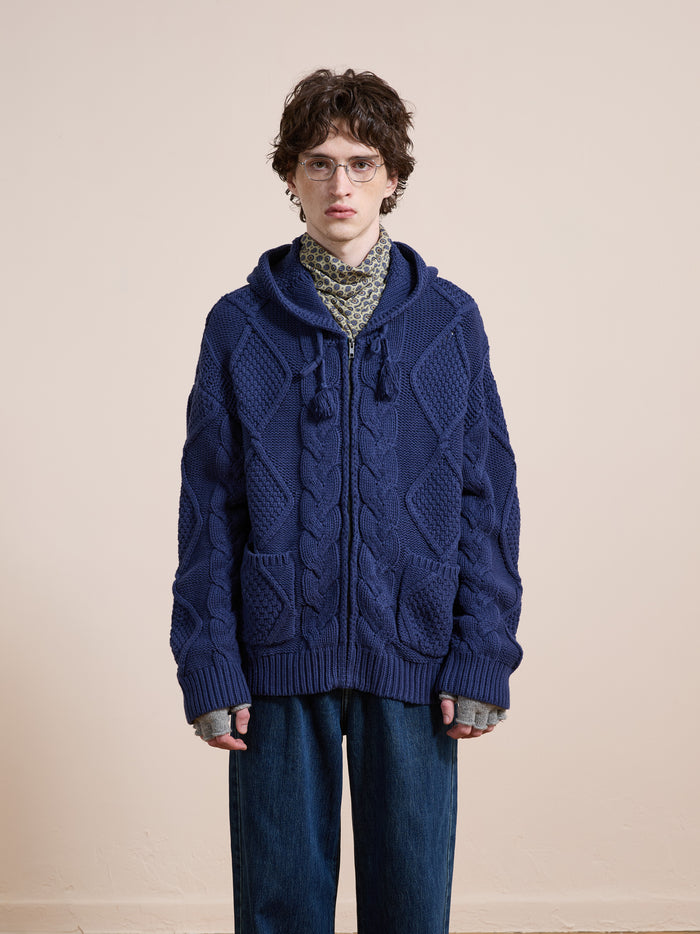 A person wearing glasses, a patterned scarf, a chunky blue Found Zip Up Cable Knit Hoodie, and loose blue jeans stands facing the camera against a plain beige background.